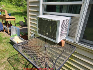 Window air conditioner unit supported by an outdoor table and bricks (C) Daniel Friedman at InspectApedia.com