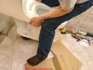 Carefully lower the new toilet and its wax seal into place straight down over the wastepipe - guiding the toilet mounting bolts through the holes in the side of the toilet base (C) Daniel Friedman at InspectApedia.com